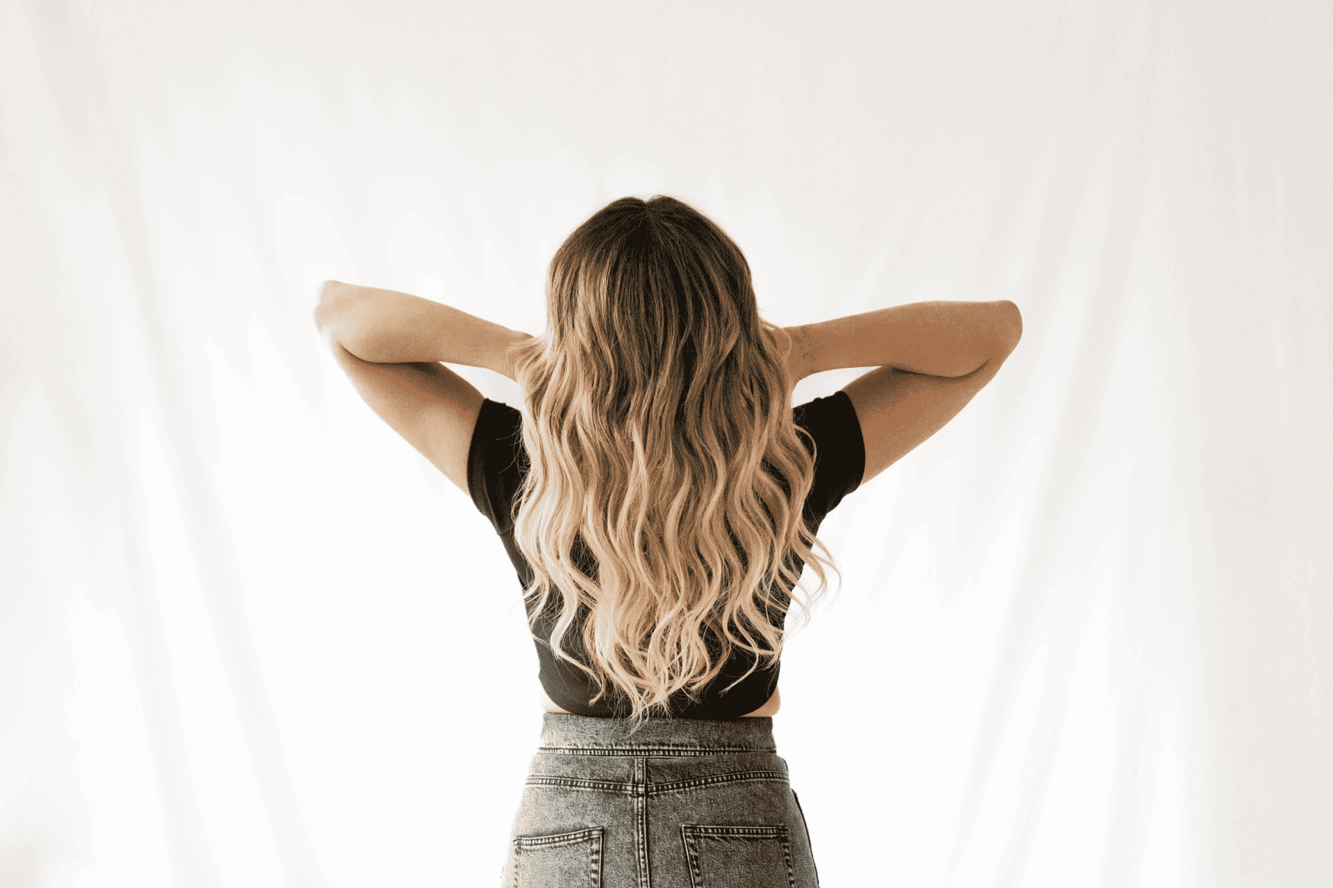Woman with long wavy hair in black top and gray skirt standing against a white background.