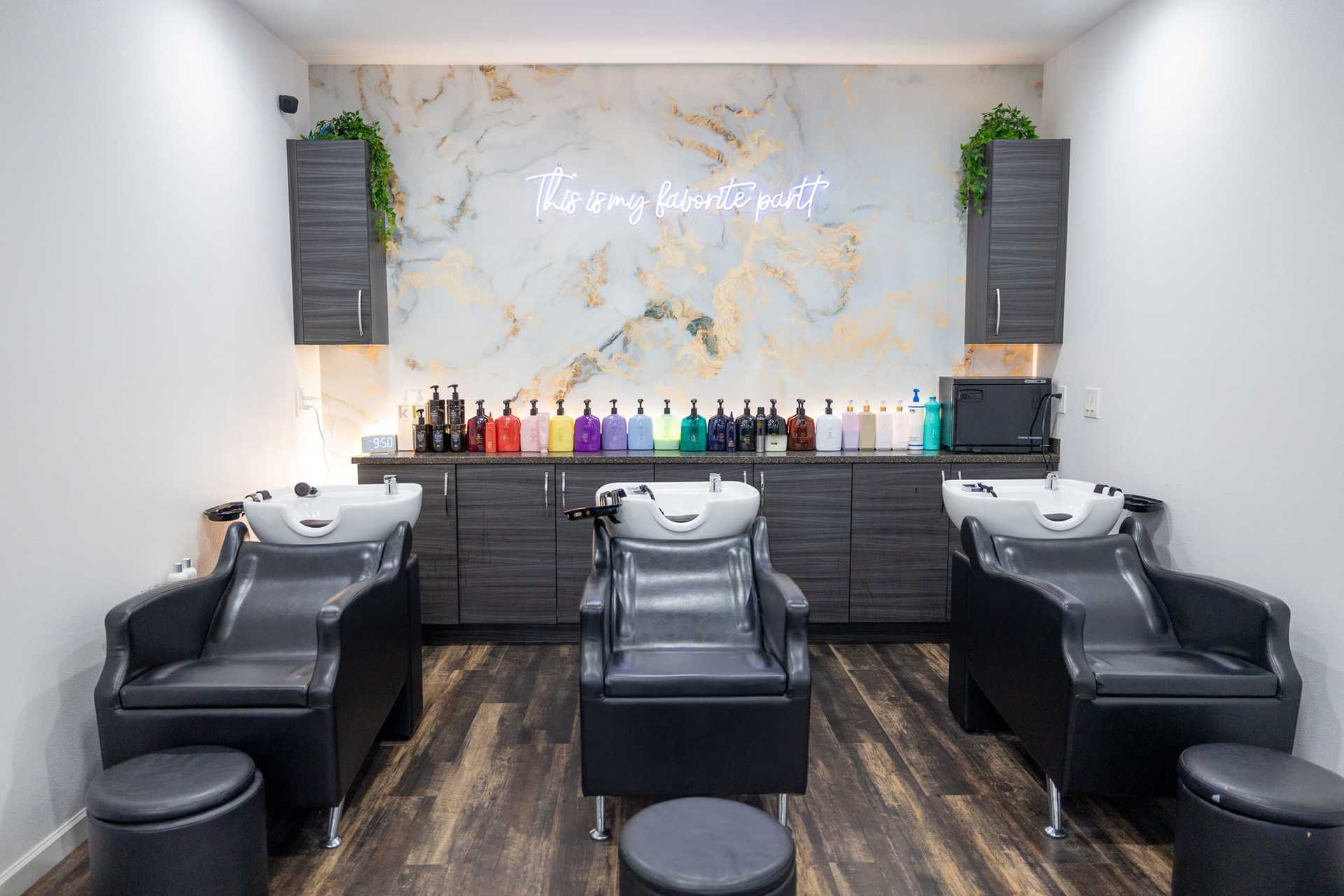 Modern salon shampoo area with black chairs, colorful bottles, and a marble wall sign.
