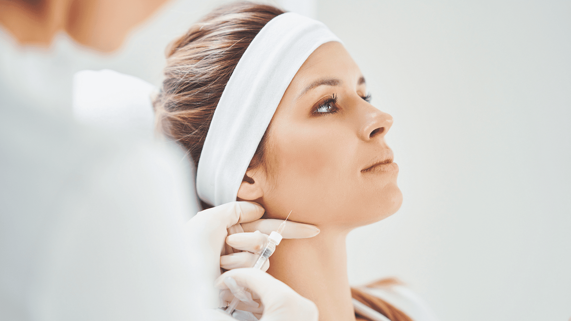 Woman receiving a cosmetic injection in a clinic, wearing a headband while a doctor administers.